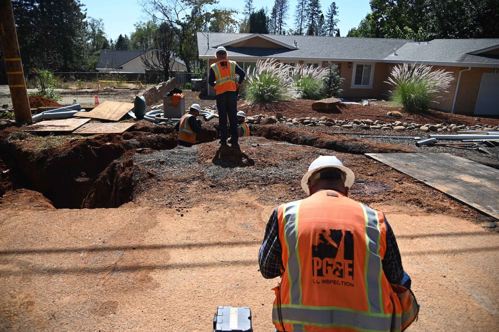 a person in an orange construction vest with PG&E on the back sits in front of a dirt path. In front of them, another worker is standing by a big hole in the dirt ground.