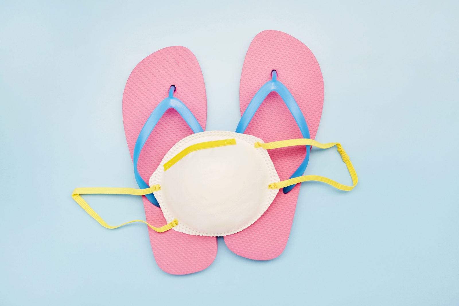 Still life of a white face mask and pink flip-flops on blue background, on vacation with a face mask