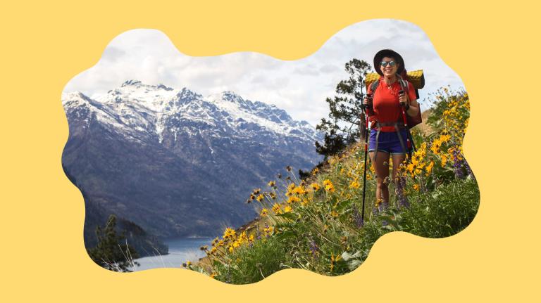 Person wearing red shirt and purple shorts stands on flower-covered mountainside with snowcapped mountains in background