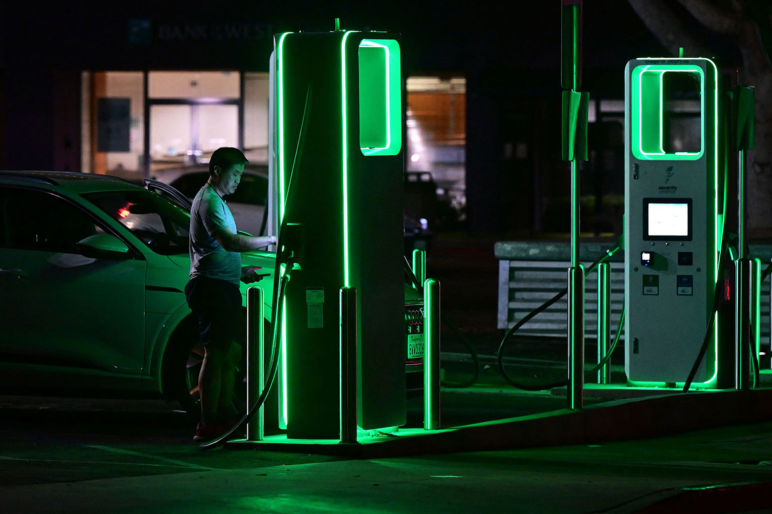 A driver, illuminated by the green, glowing charging station, charges their electric vehicle