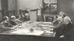 Man in a waistcoat talking to a committee of similarly dressed old white men