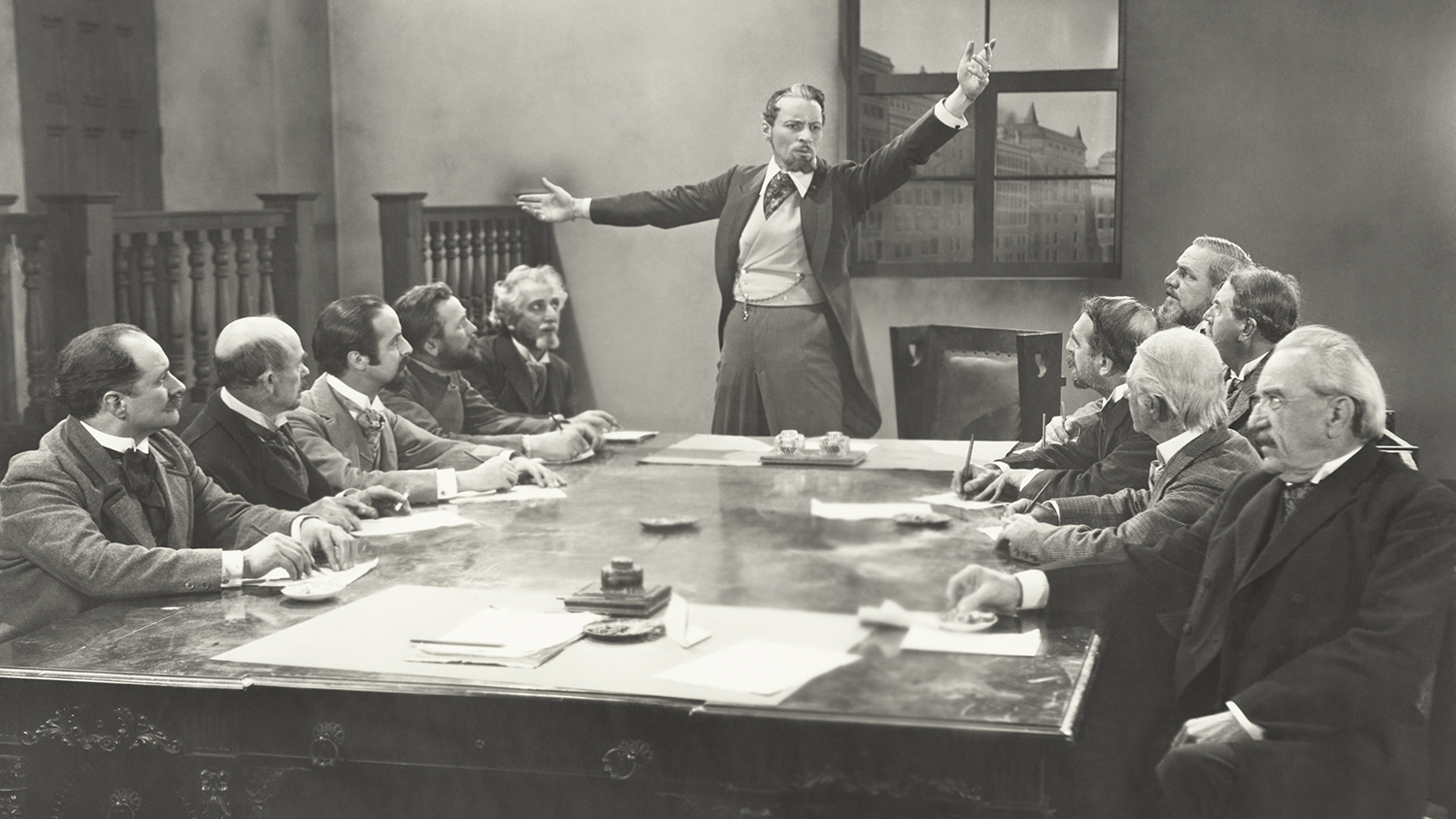 Man in a waistcoat talking to a committee of similarly dressed old white men