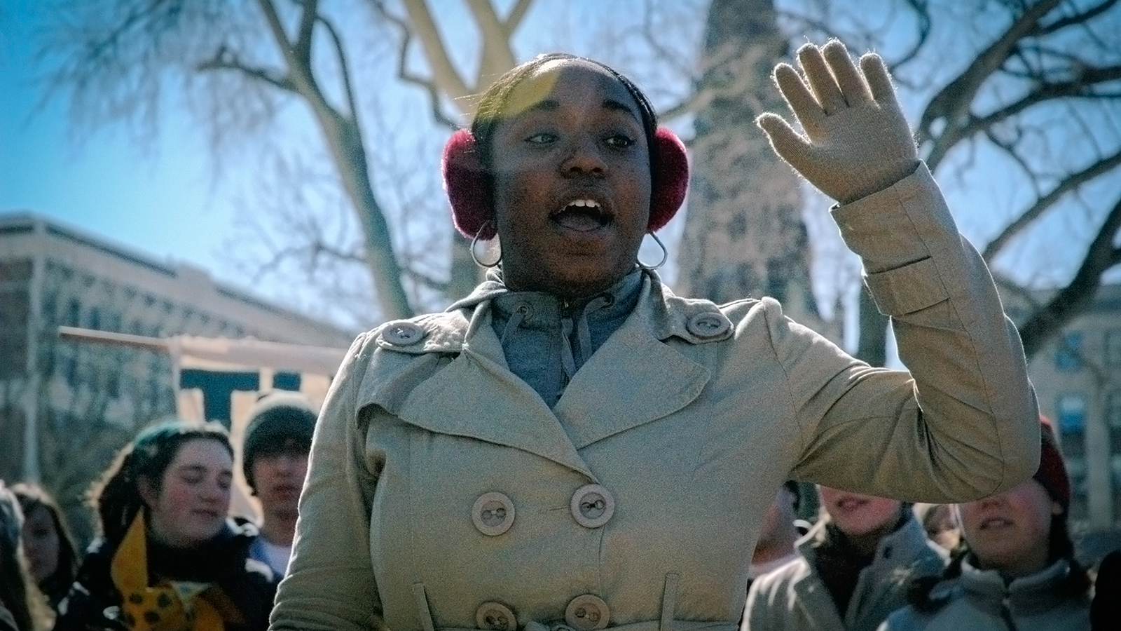 A woman in red earmuffs speaks with one raised against a blue sky with people and trees in the background