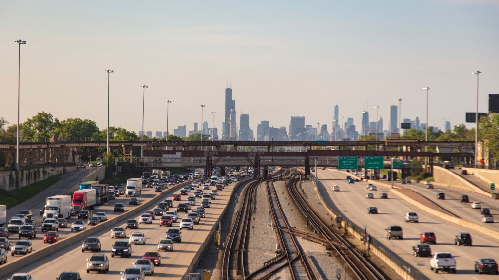 Elevated view of train tracks and multiple freeway lanes leading to Chicago. The skyline of Chicago covers the horizon. Moderate traffic leading into the city and heavy traffic leading away.