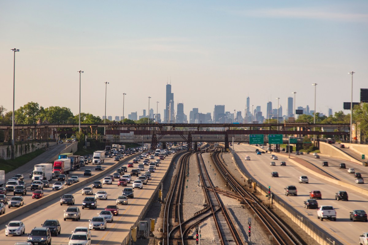 Elevated view of train tracks and multiple freeway lanes leading to Chicago. The skyline of Chicago covers the horizon. Moderate traffic leading into the city and heavy traffic leading away.