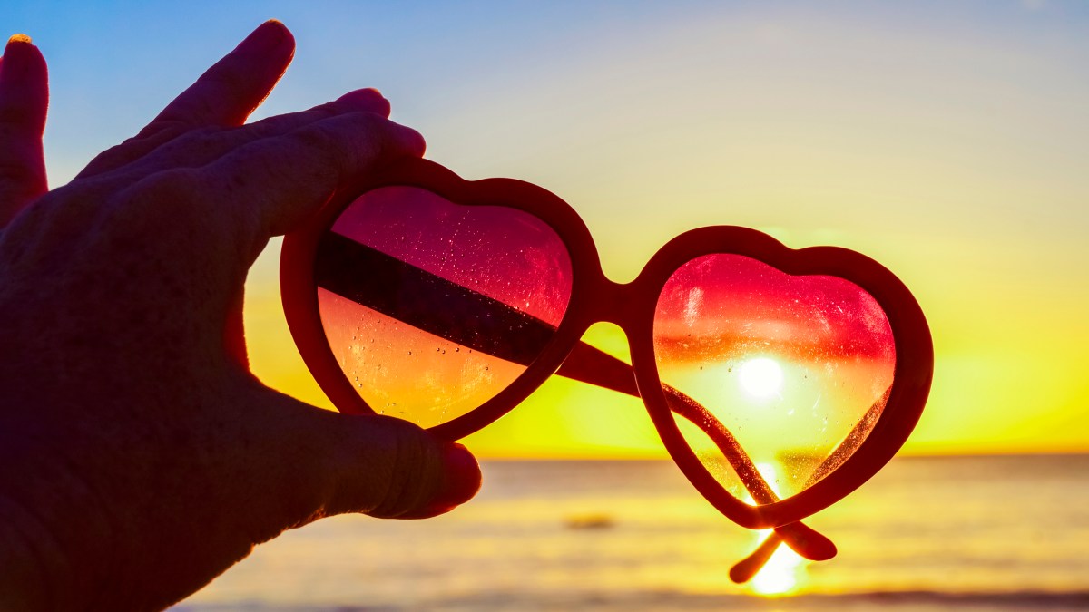 Hand holding heart-shaped sunglasses against the sun at beach, summer day
