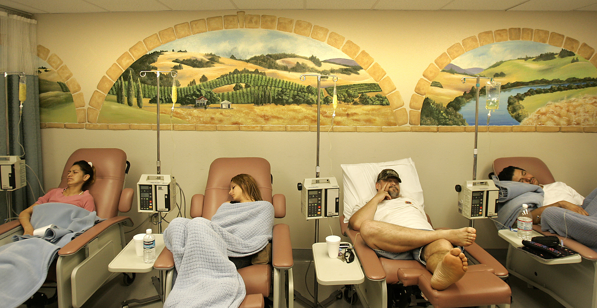 Four medical beds are set close to each other each one with a patient looking sick. Behind them, a series of murals of California