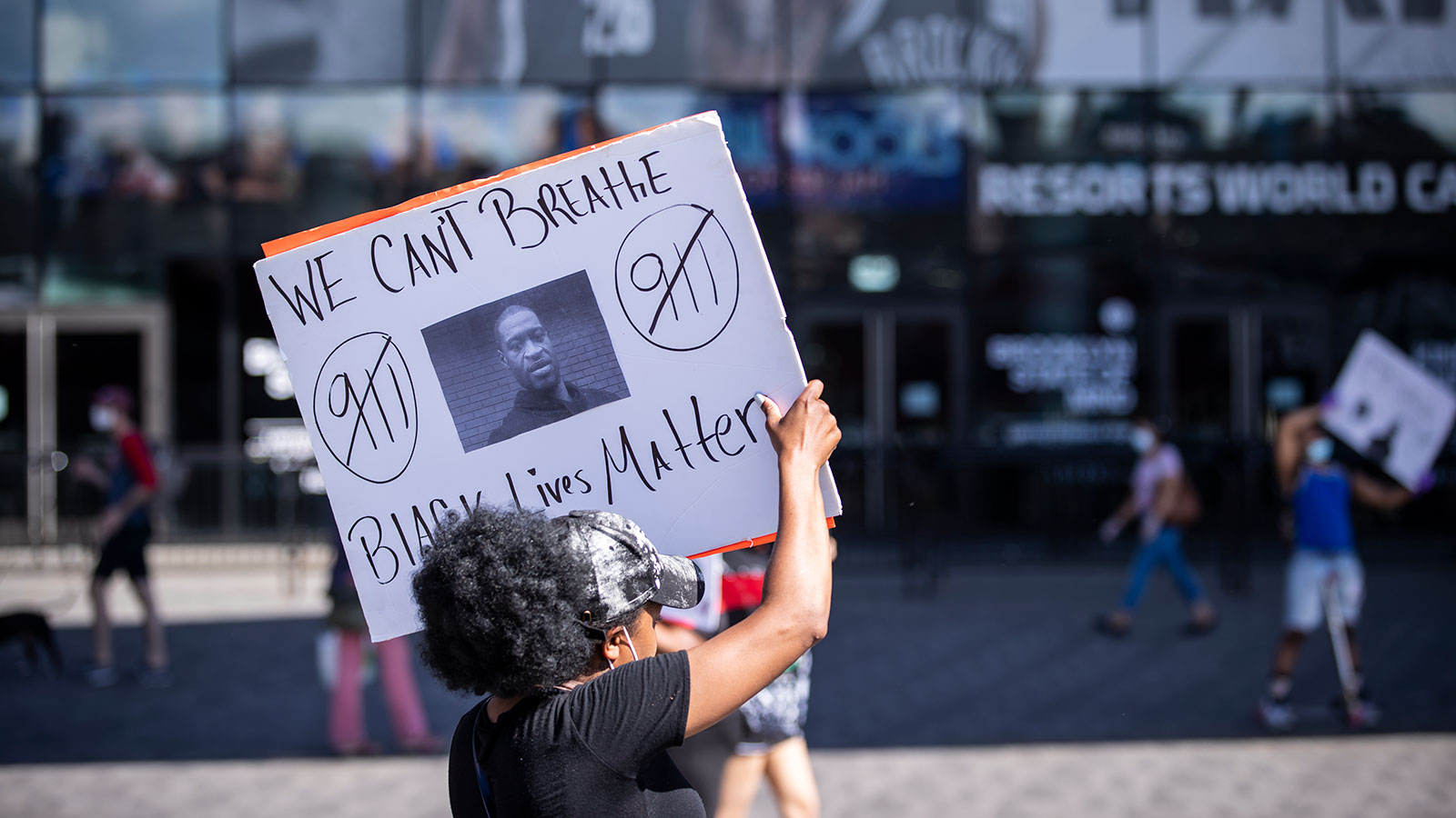 A protester wearing a mask around their neck holds a sign with a picture of George Floyd and saying We Cant Breathe and Black Lives Matter.