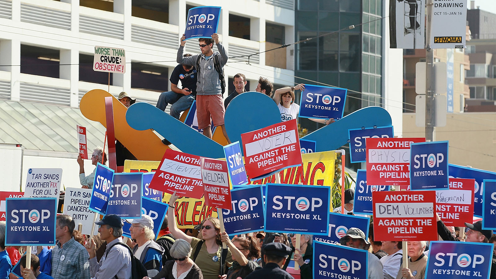 Protestors against the construction of the Keystone XL oil pipeline hold signs and stand on a Keith Haring sculpture as they demonstrate outside of the W Hotel before the arrival of U.S. President Barack Obama on October 25, 2011 in San Francisco, California