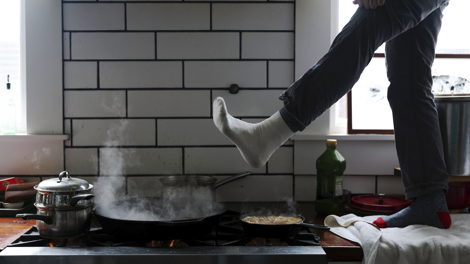 A person stands on a kitchen counter with their foot extended over their gas stove for warmth