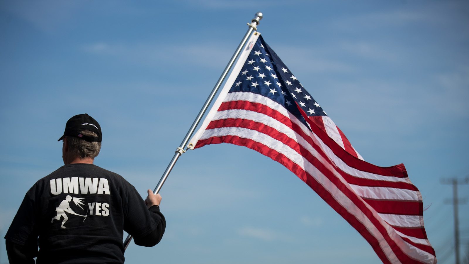 A coal miners' union member in a black shirt with white writing on the back holds an American flag before the start of a campaign rally with United Mine Workers of America.