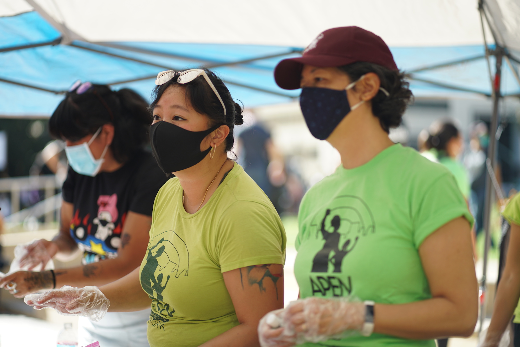 Two people in black face masks and green APEN t-shirts look to the left on the camera. They are wearing plastic gloves on their hands.