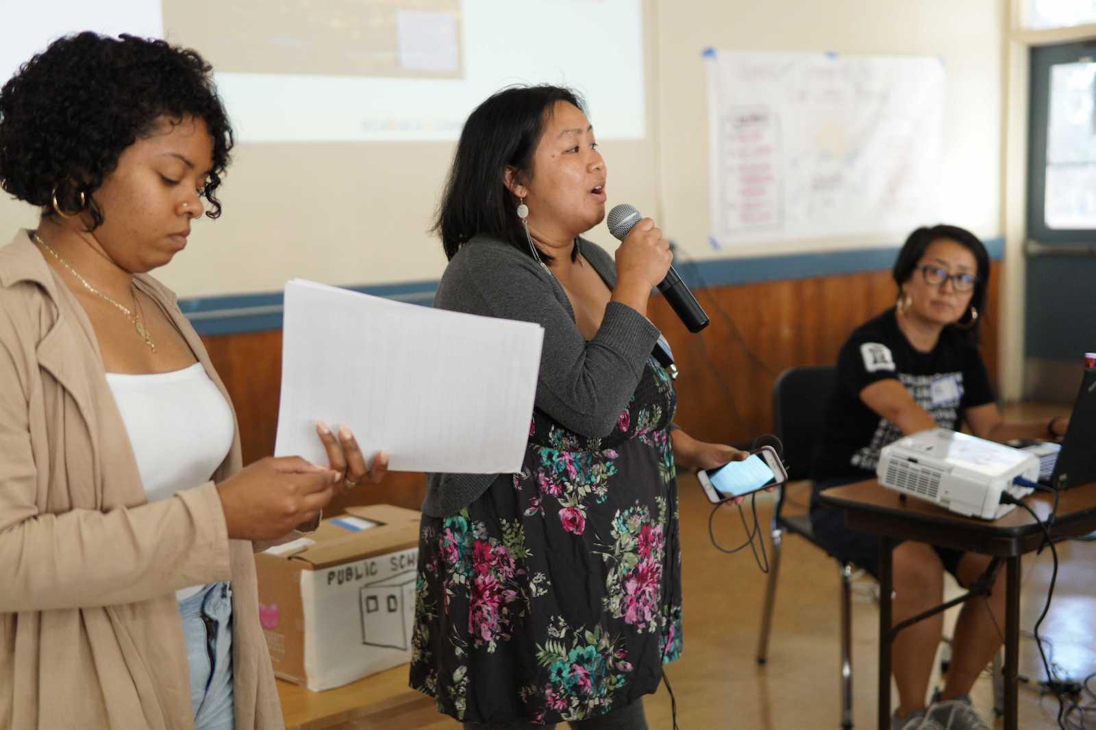 A woman in the center of the photo holds a mic and a cell phone while speaking at a community event. She is flanked by two other women who appear to be listening to her.