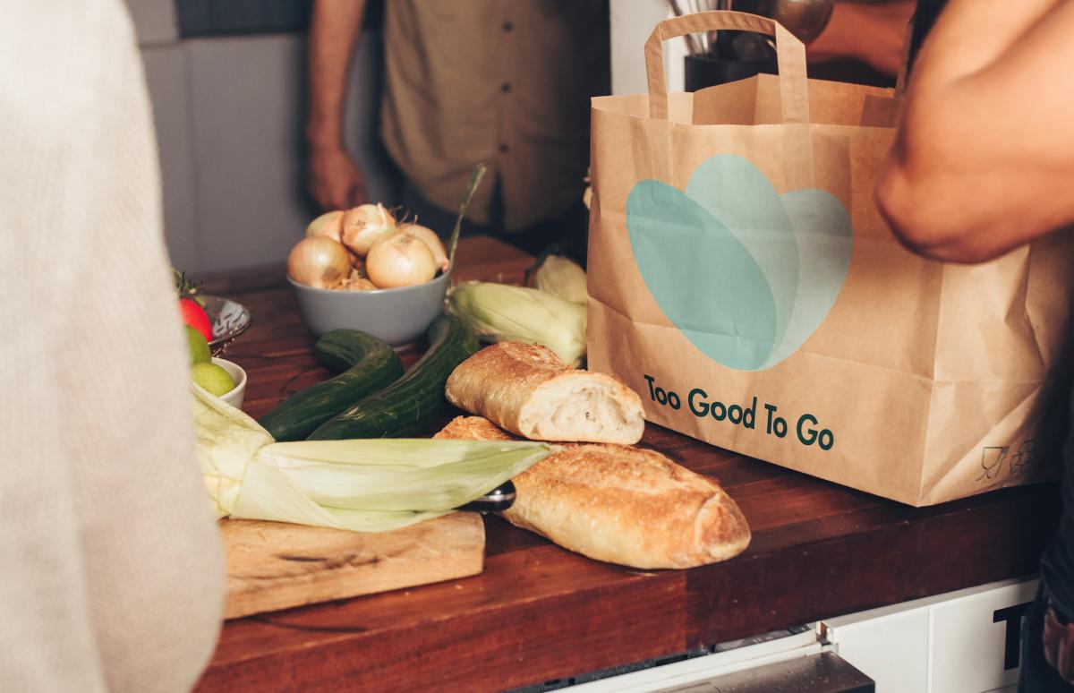 A photo of bread, onions, and zucchini on a wooden table (left) and a large brown paper bag with three blue circles and the words "Too Good To Go" (left)