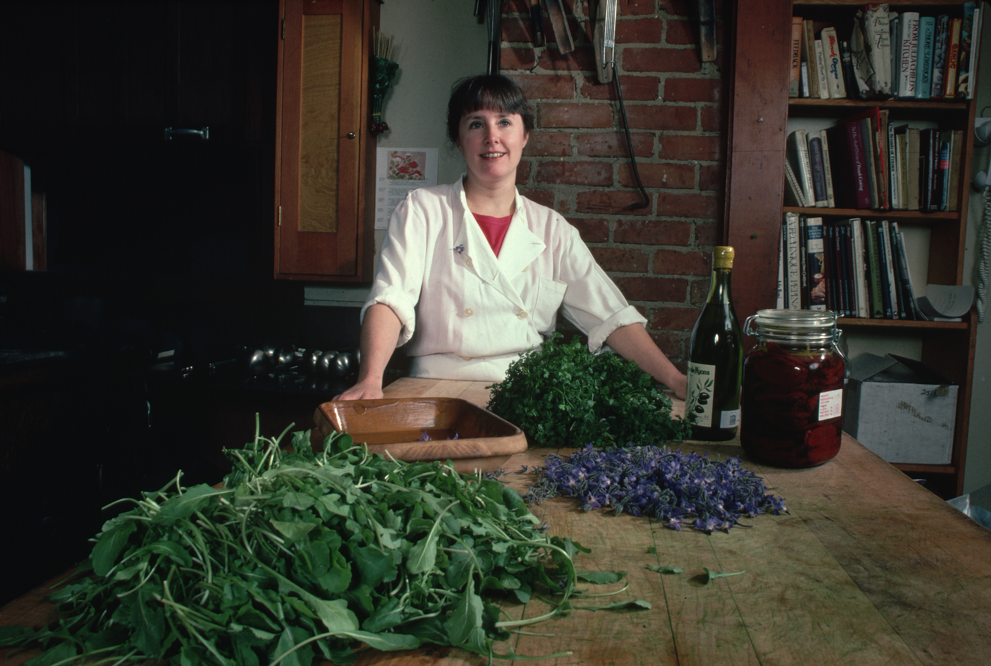 Chef Alice Waters stands behind a large wooden table covered in fresh produce, a large pile of baby greens on the left.