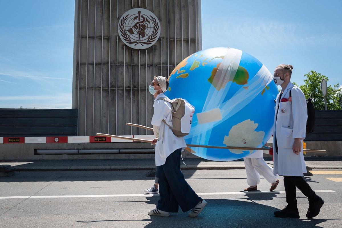 two people in white medical coats carry a blow up globe covered in bandages as they walk in front of a building with a medical logo