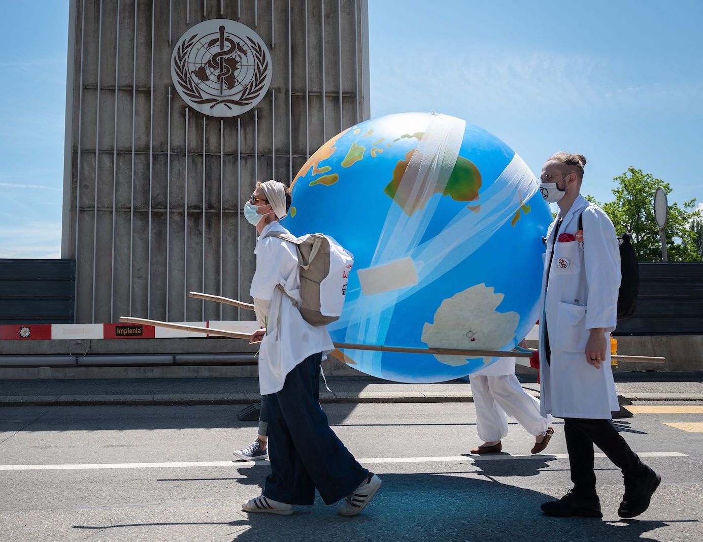 two people in white medical coats carry a blow up globe covered in bandages as they walk in front of a building with a medical logo