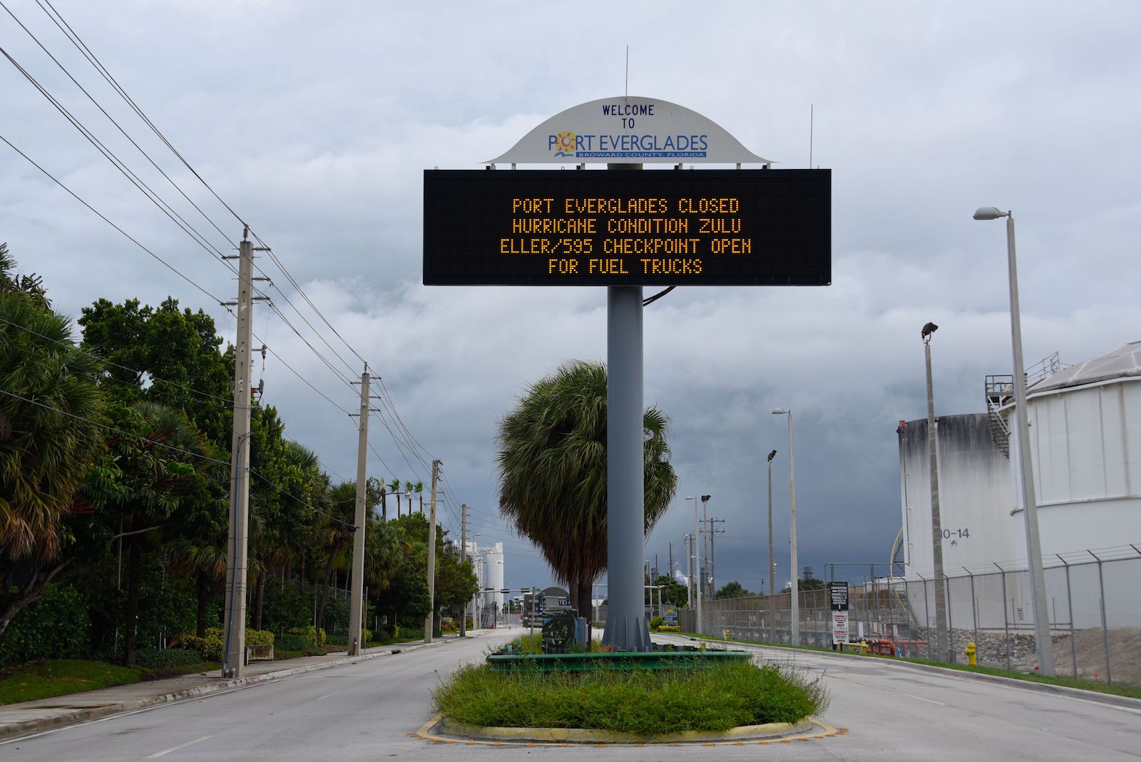a large, tall, metal Port of Everglades sign with a black backround and orange light-up lettering stands at the center of the image. It reads, "Port Everglades closed. Hurricane condition zulu, eller/595 checkpoint open for fuel trucks." Behind the sign, the sky is dark and stormy.