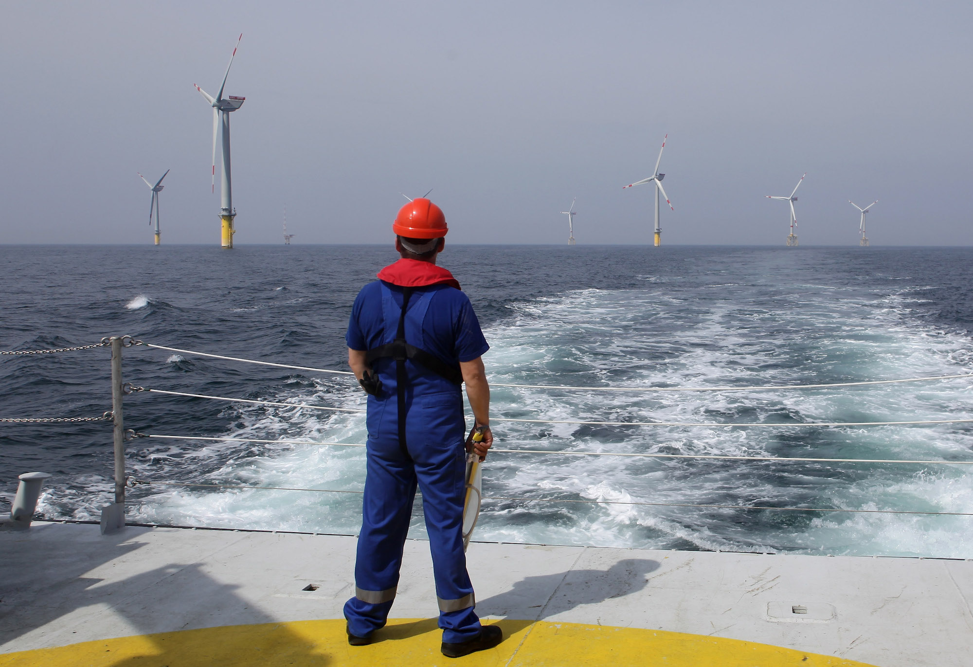 a worker in a dark blue jumpsuit and red hard hat stands on a platform looking out at several offshore wind turbines