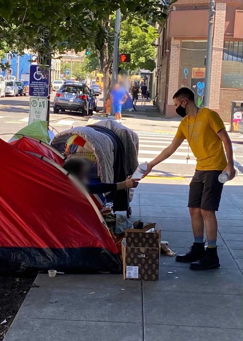 a person in a yellow shirt hands a bottle of frosty water to a person in a red tent on the streets of portland
