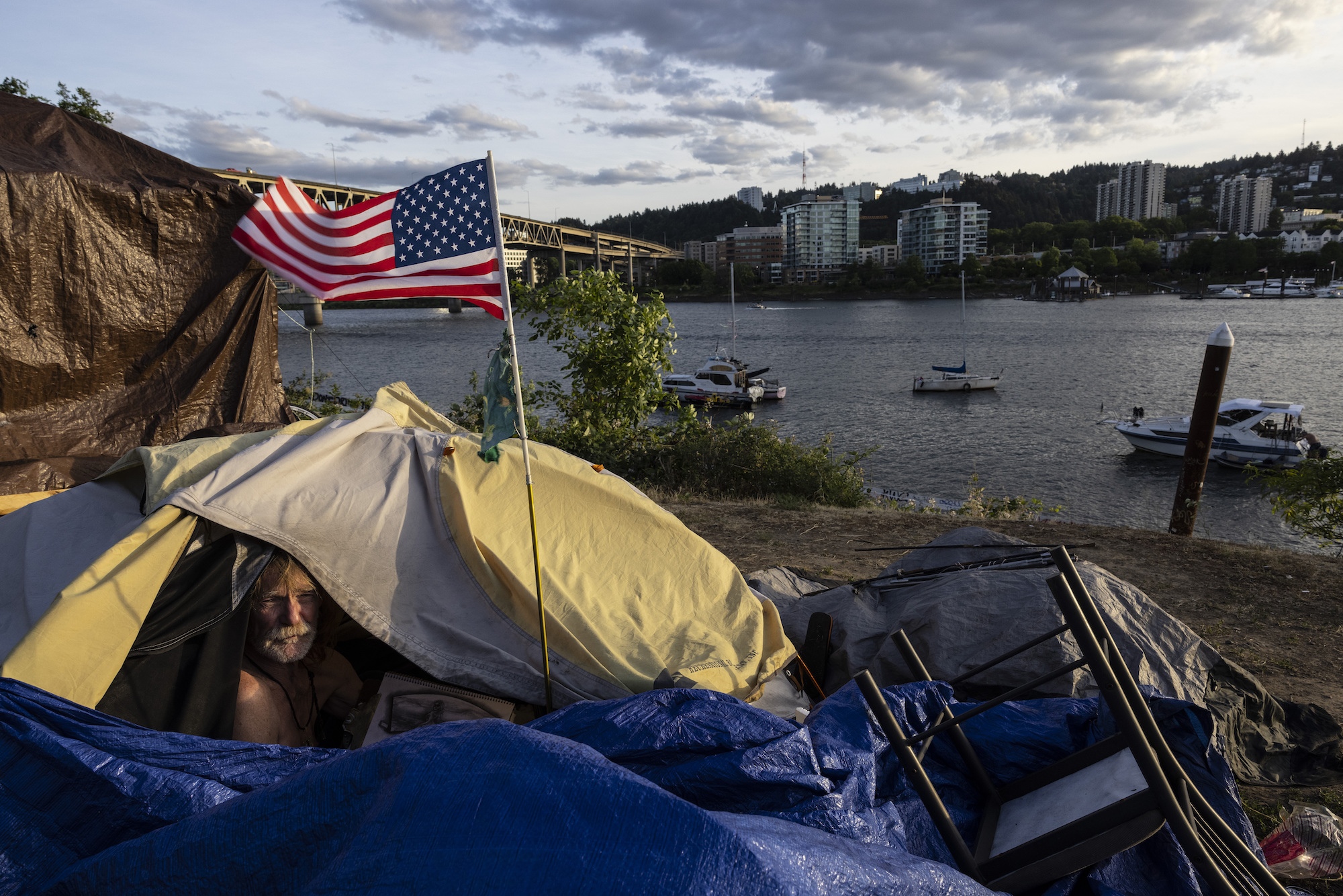 a bearded man in a yellow tent with blue tarp looks out of the flap. Outside of the tent is an American flag. The tent is on a beach with houses in the background and boats in the water.