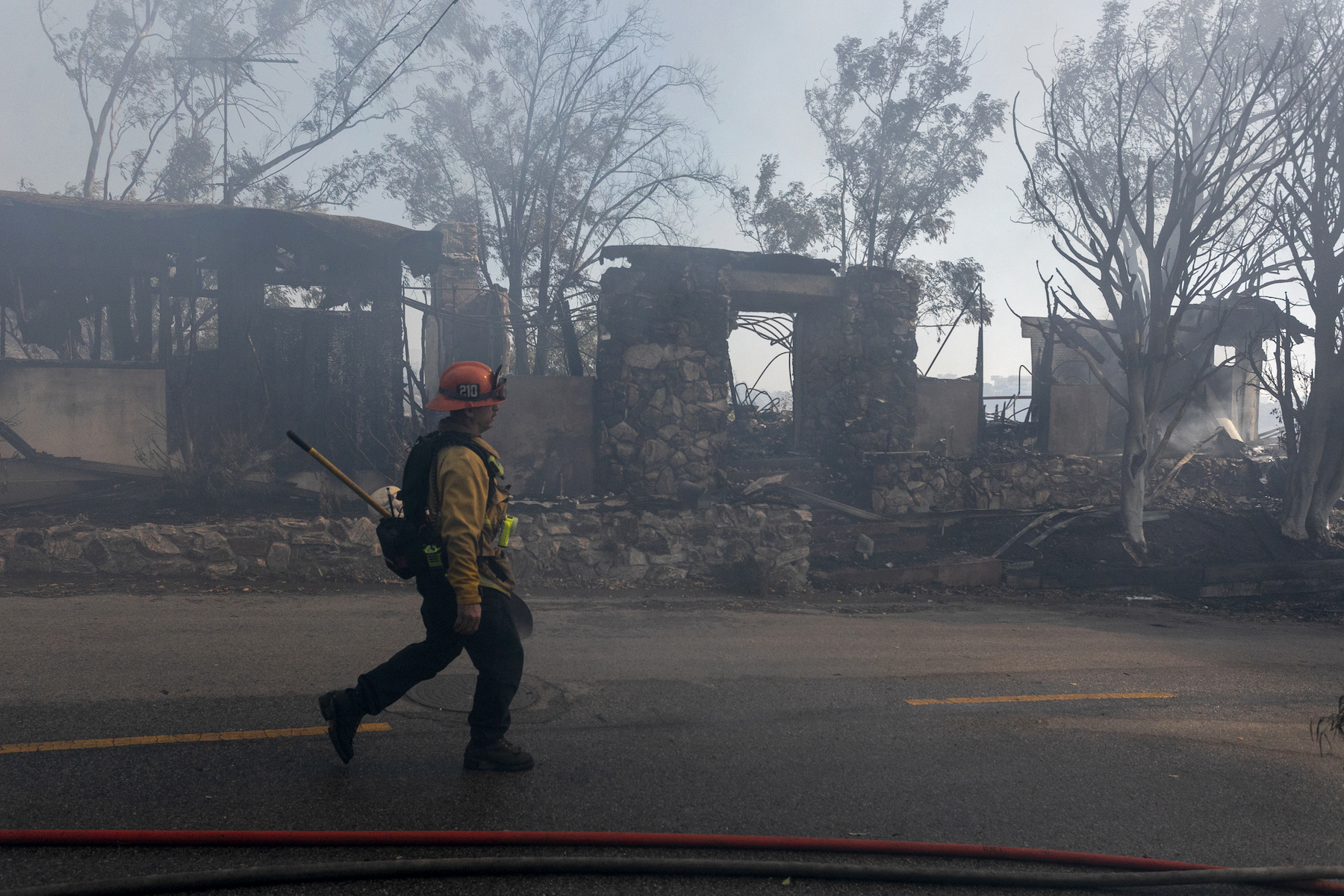 A firefighter wearing a yellow vest, orange helmet and black pants walks past burned houses in Los Angeles. In the background, against the blue sky, semi-burnt trees are visible.