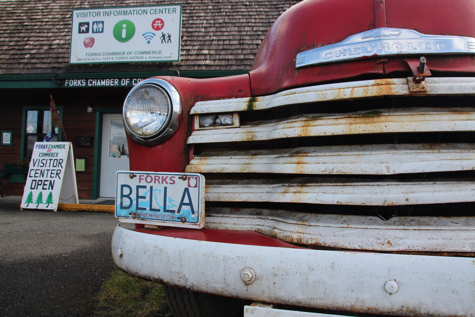 An old red truck with a dirty white grate and a license plate that says BELLA is parked in front of a building with a sign marked visitor's center