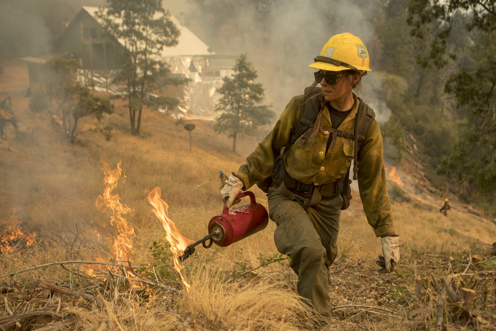 a wildland firefighter in a yellow hat and fire gear holds a red can as she walks through a dry field during a controlled burn