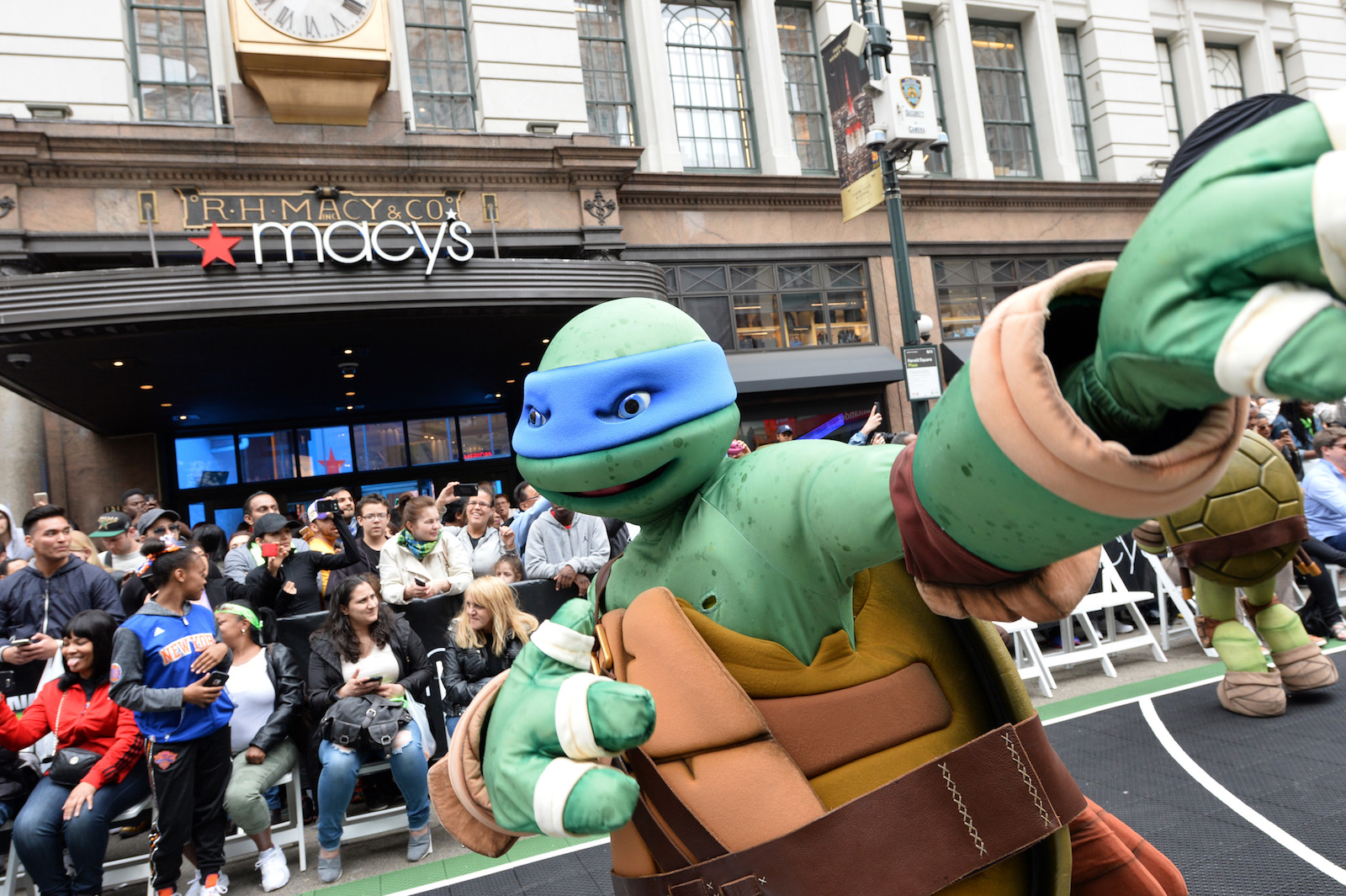 a person in a teenage mutant ninja turtle costume points into the crowd in front of a macy's department store