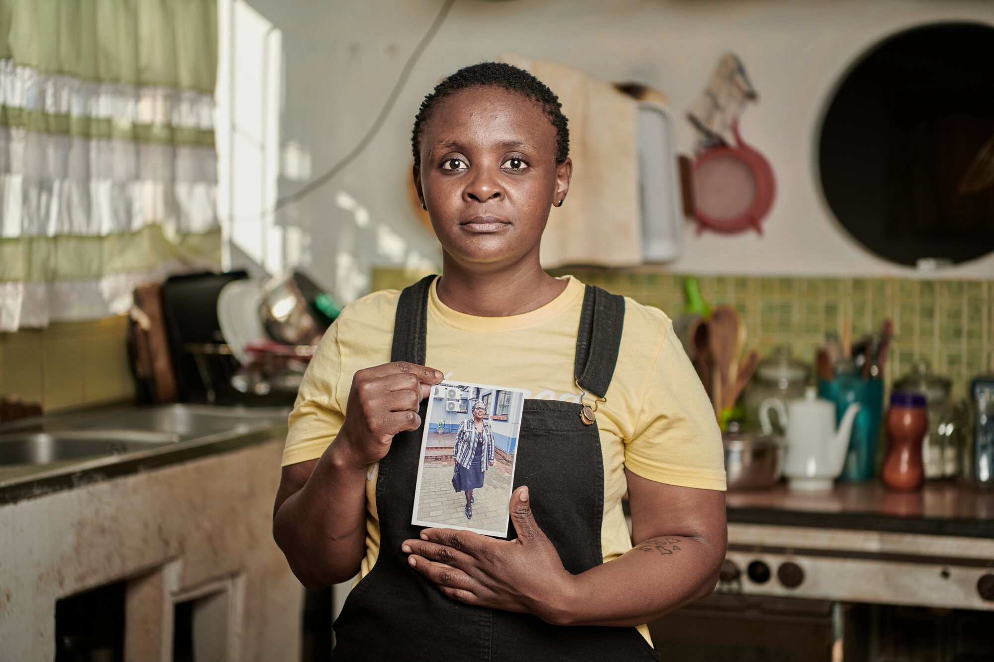 Awoman wearing a light yellow t-shirt and navy blue overalls holds a picture of her mother, an older woman. In the background you can see a kitchen with light beige walls and lime green tiles.