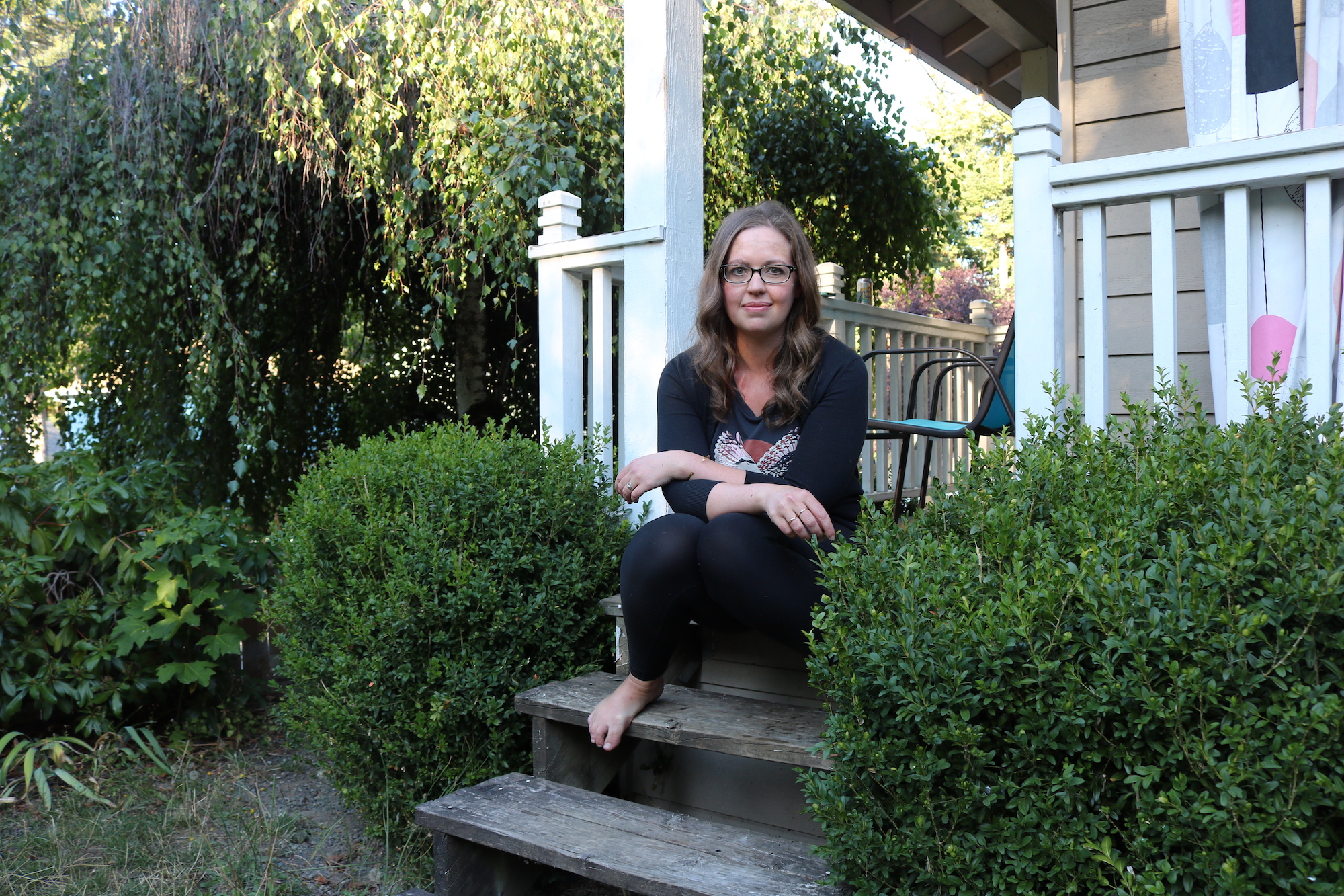 a woman sits on the steps of a houe with white trim surrounded by green bushes
