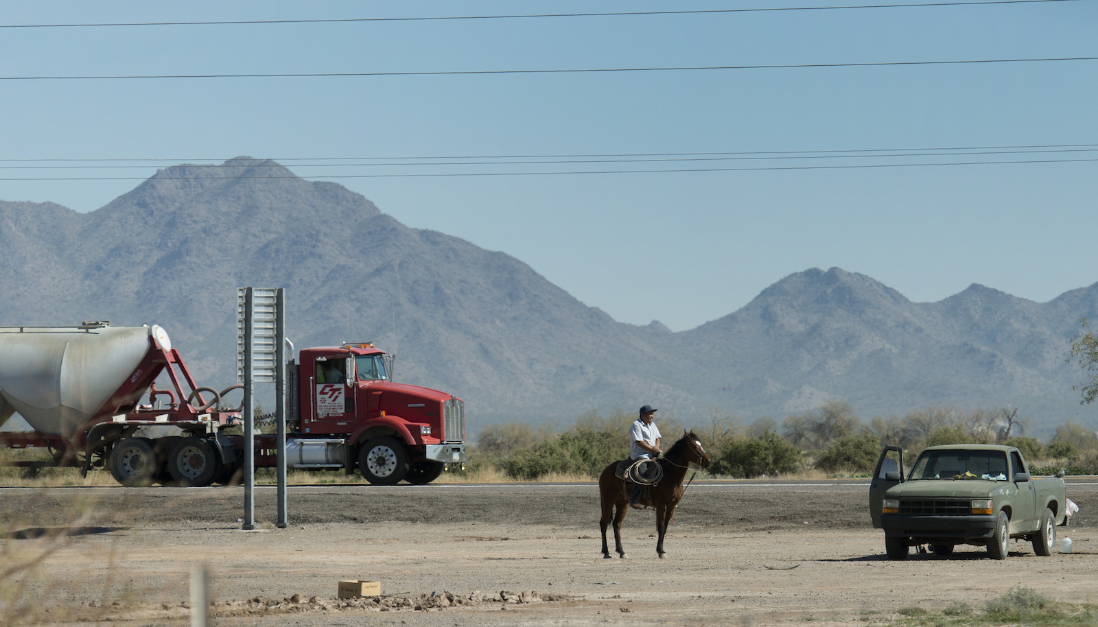 a red truck, a man on a horse, and a pickup truck stand on a dry, mountainous landscape