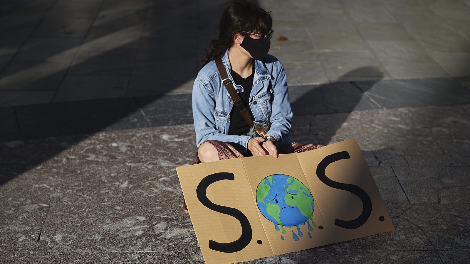 A girl sits on the ground alone with a sign saying SOS and a sad-looking Earth.