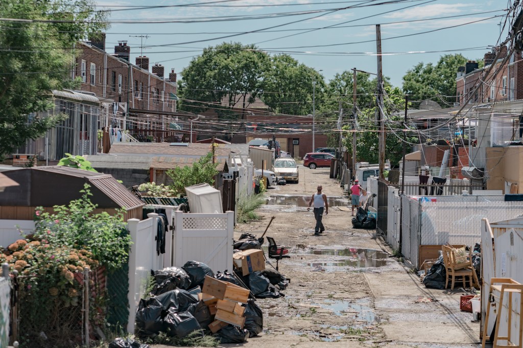 A street in New York looks wet, garbage and debris acoomultes in the sidewalk. A man wearing a white tank top and jeans walks through it.