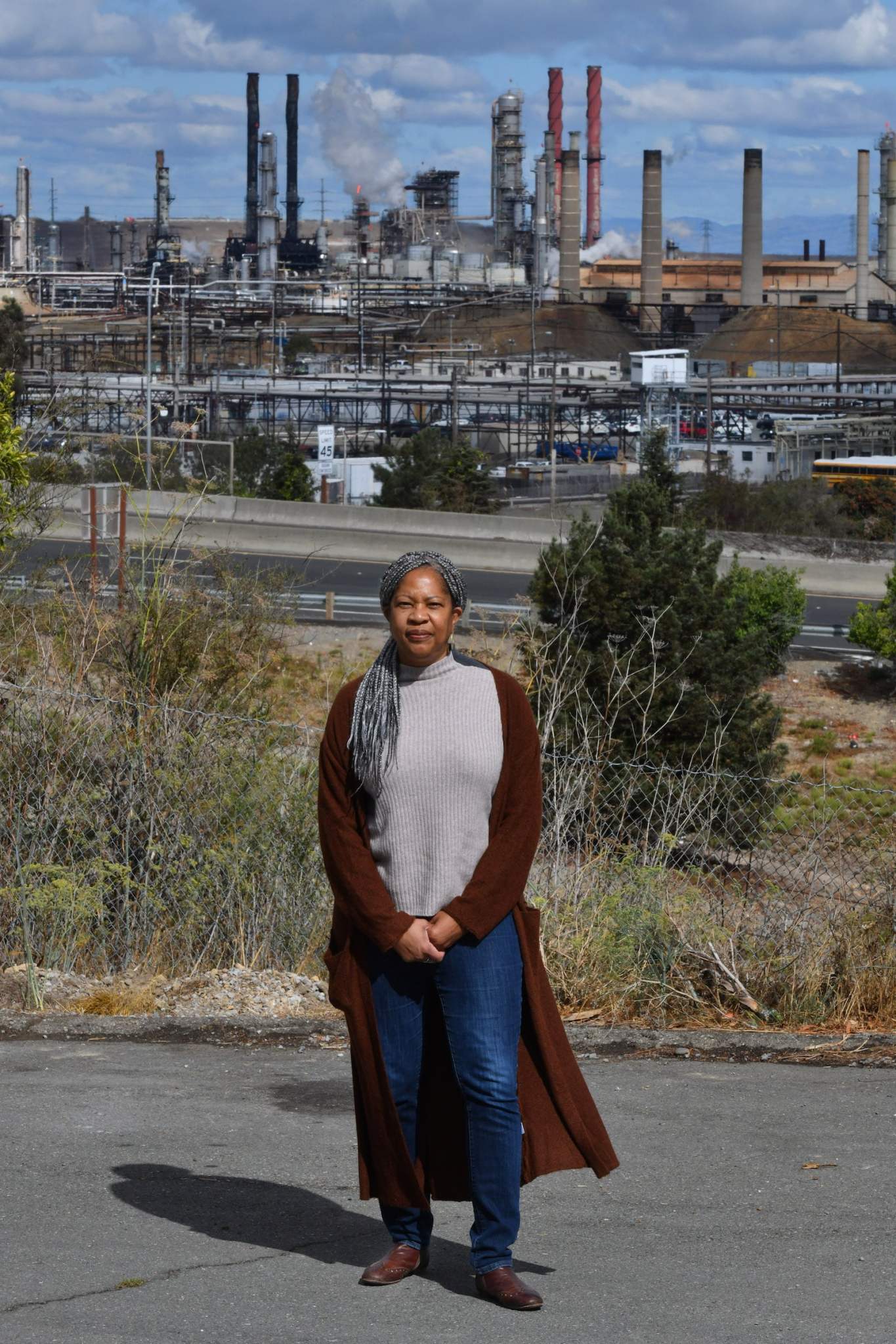 a woman in a gray shirt and red long coat stands in front of a refinery