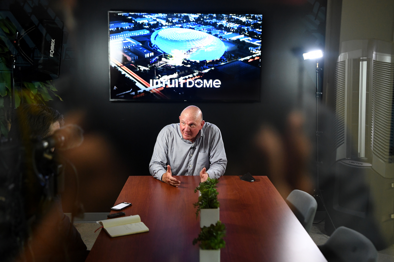 an man in a gray shirt sits at the end of a wooden conference table with a tv showing a stadium interior behind him