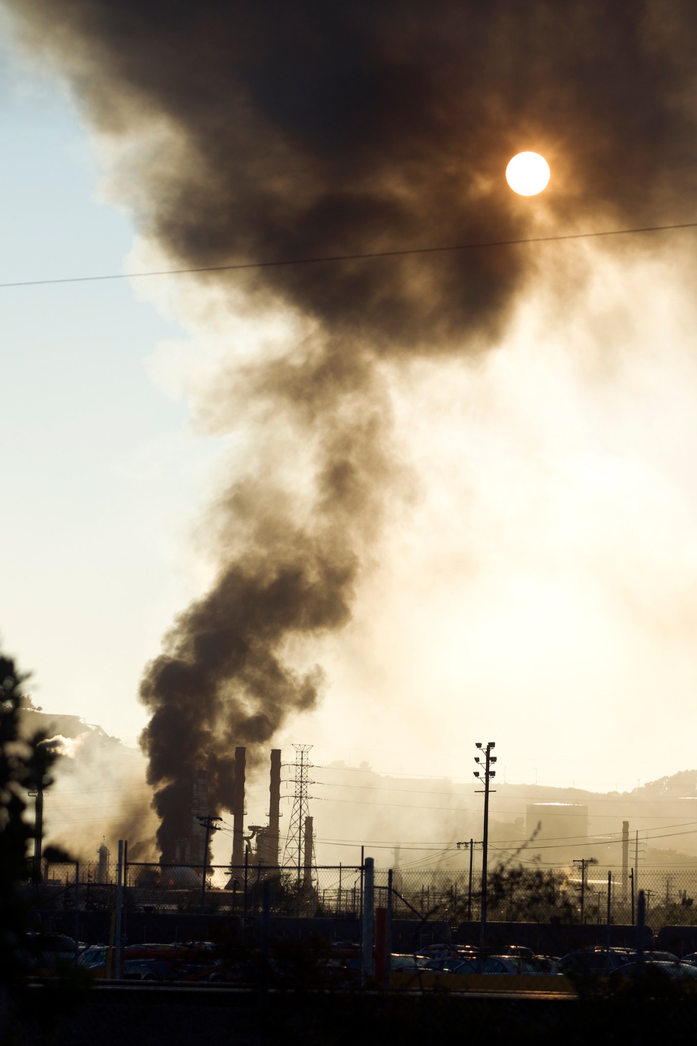 a large cloud of smoke from a silhouetted refinery reaches high into the sky partially obscuring the sun