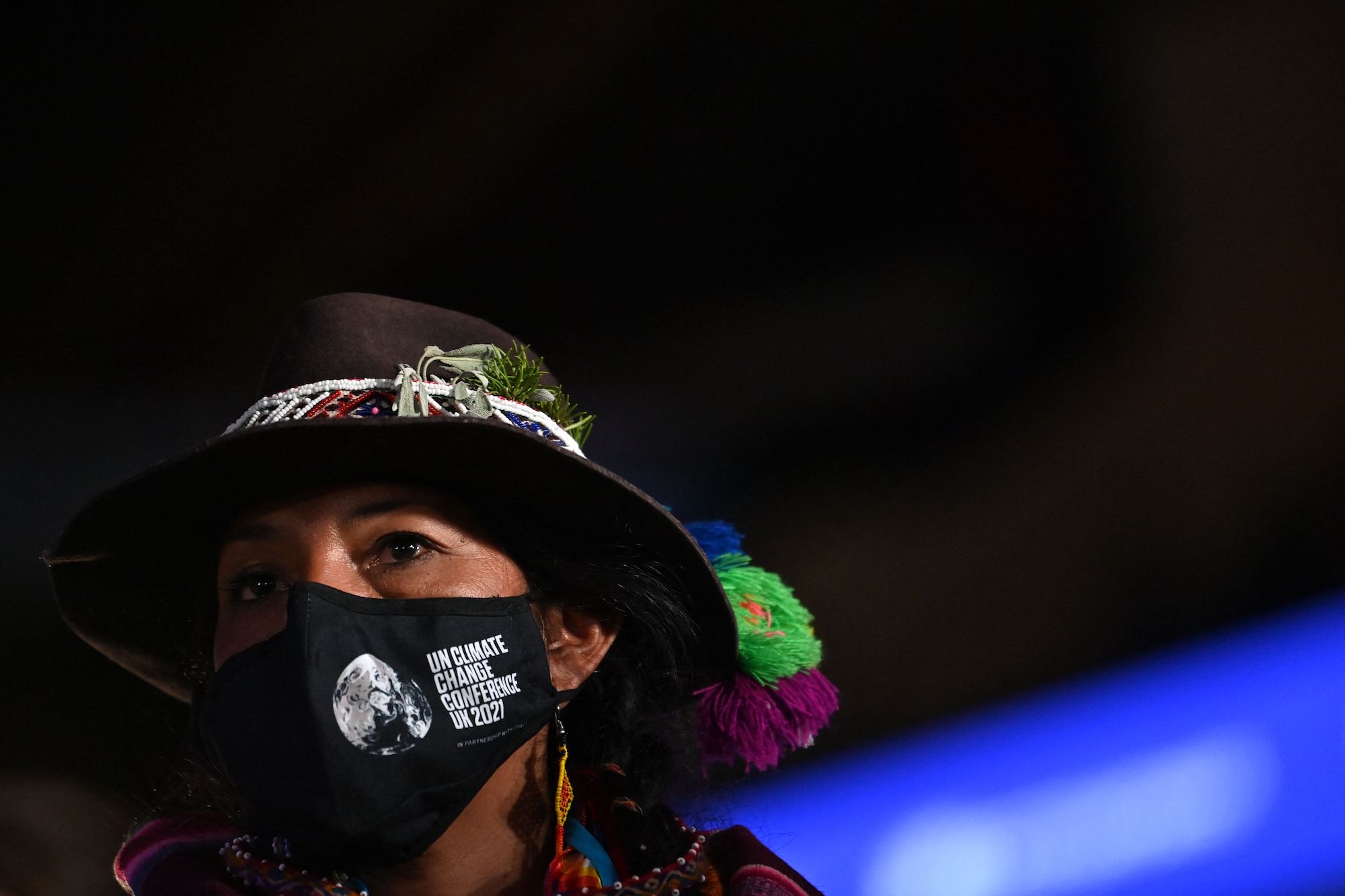 A close up photograph of a woman wearing a traditional quichua hat and a mask with the words 
