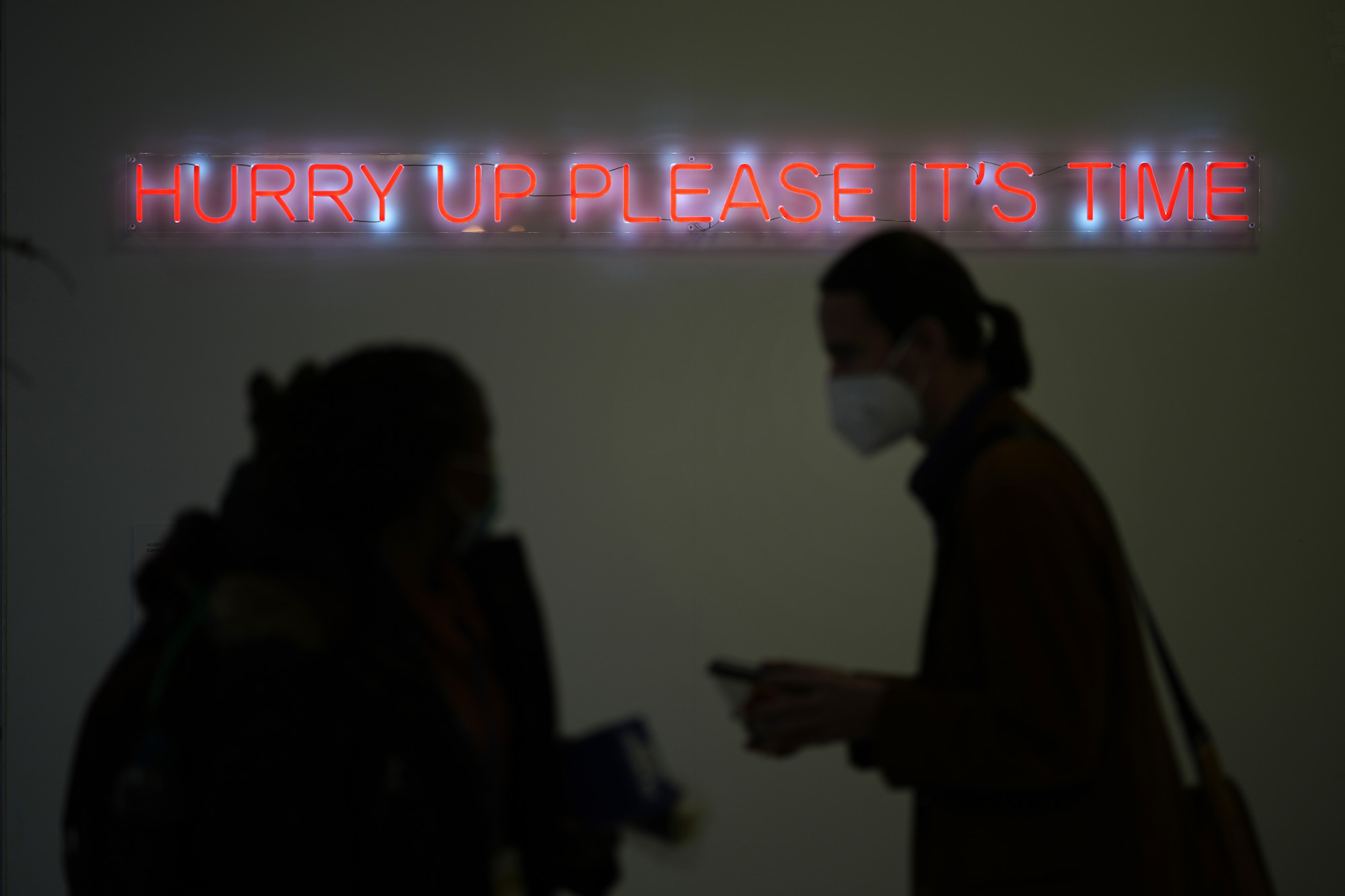 silhouettes of people walk past a neon sign that says hurry up please it's time