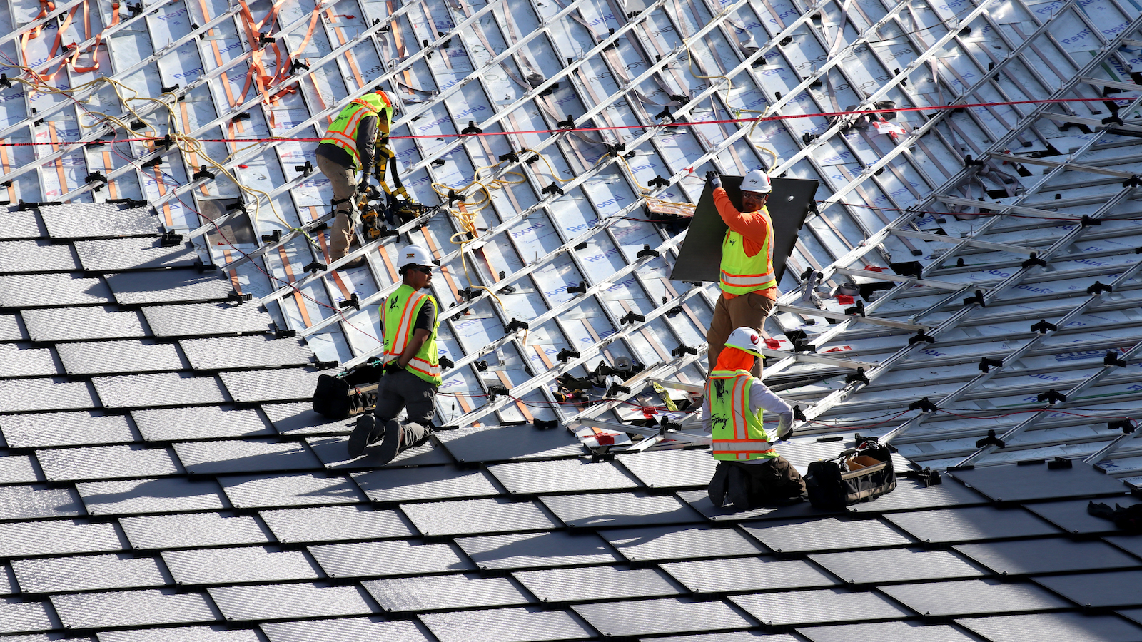 people in construction vests and hard hats kneel or stand on a roof patterned with overlapping diamond-shaped solar panels like scales