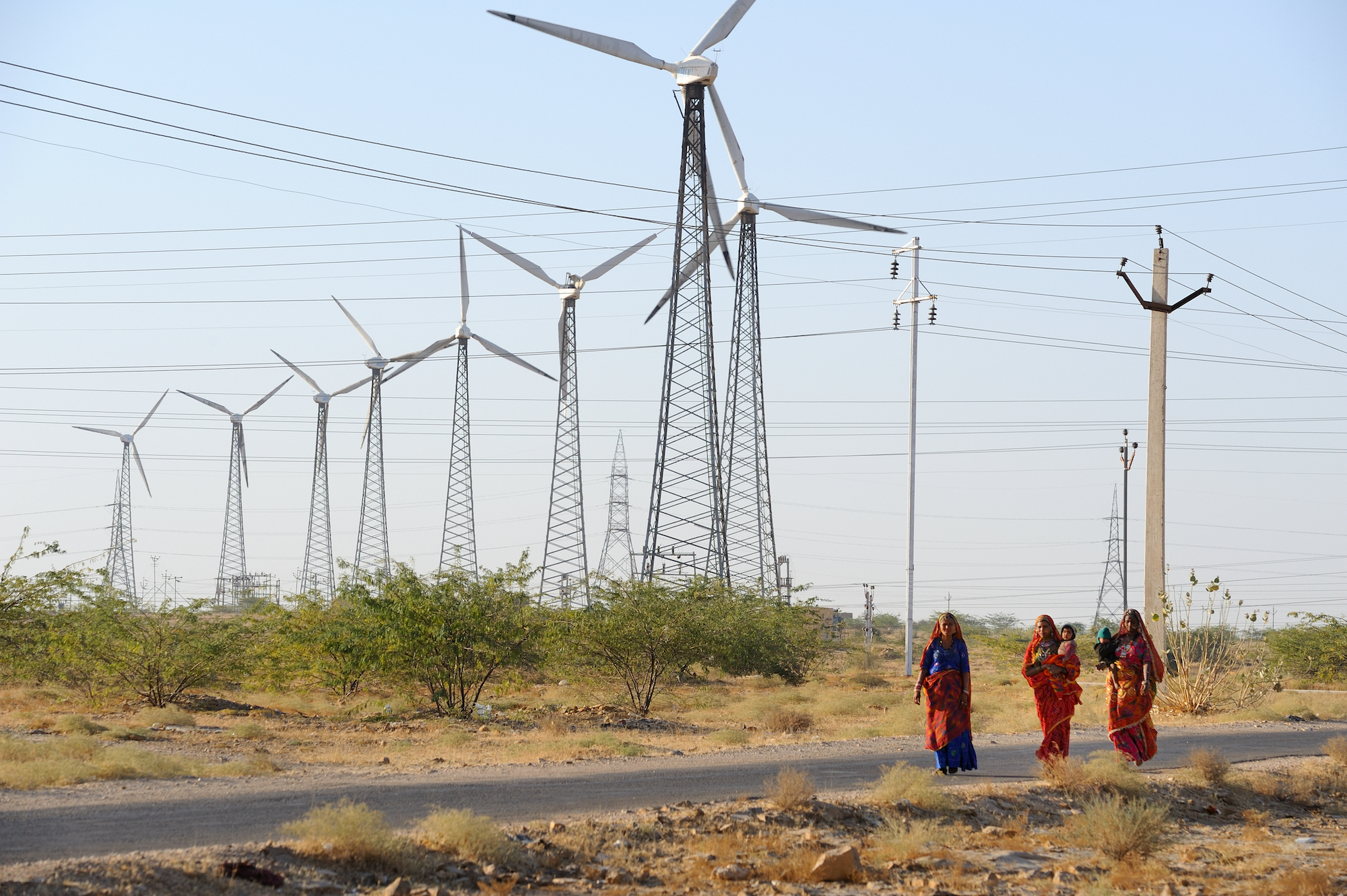 There women using red and bule saris walk on a pavement road in india. on the background, against a pale blue sky, several wind energy mills sprout from the earth.