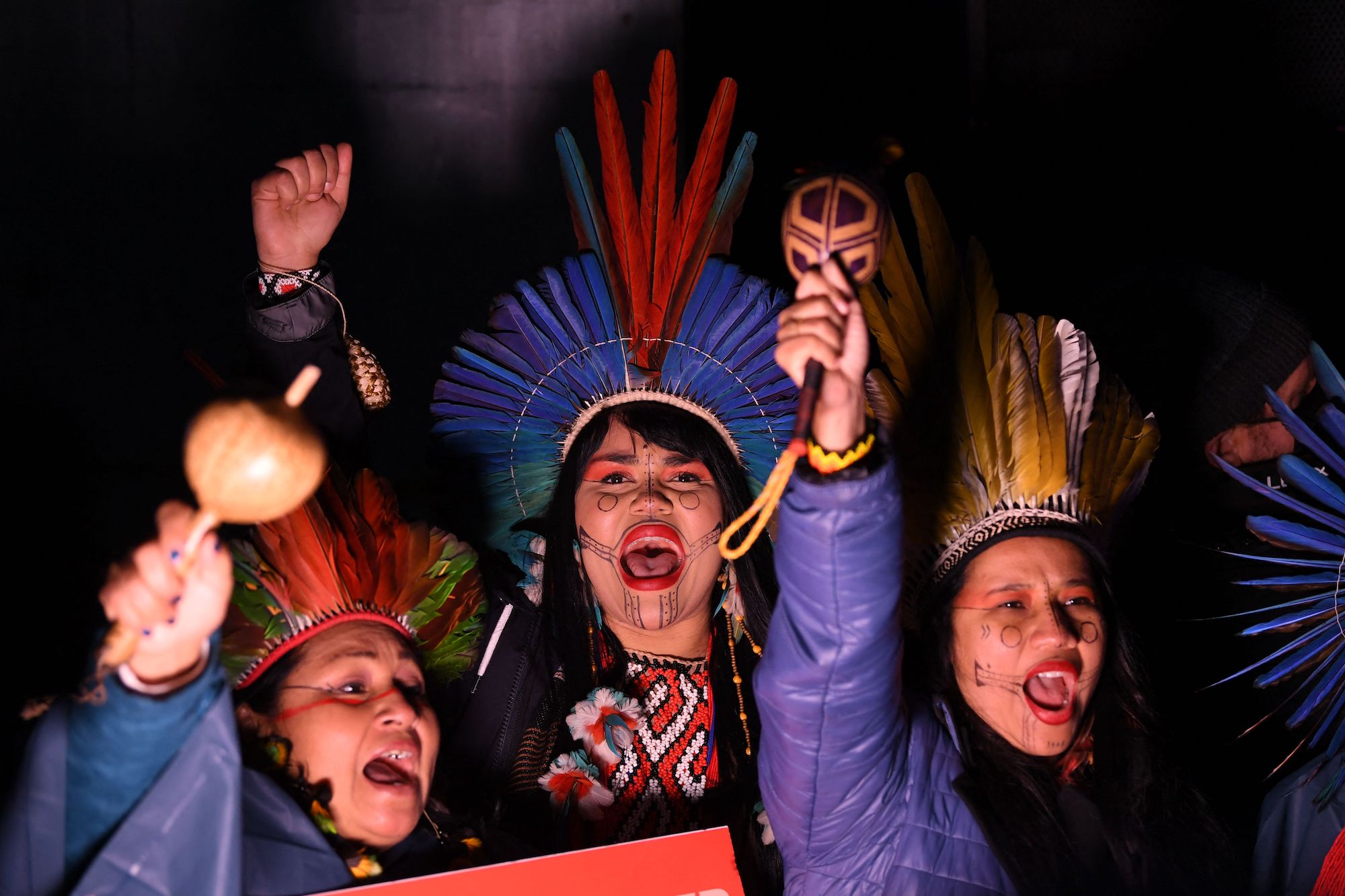 Three Indigenous women with feather crowns and their faces painted chant, raising their hands.