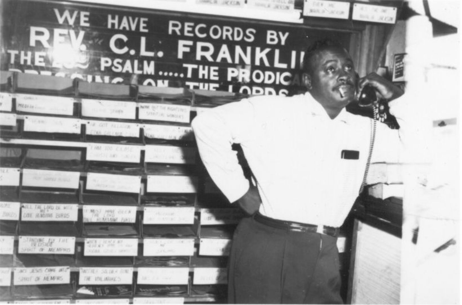 a man in a black and white photo leans against a tall stand in a record store