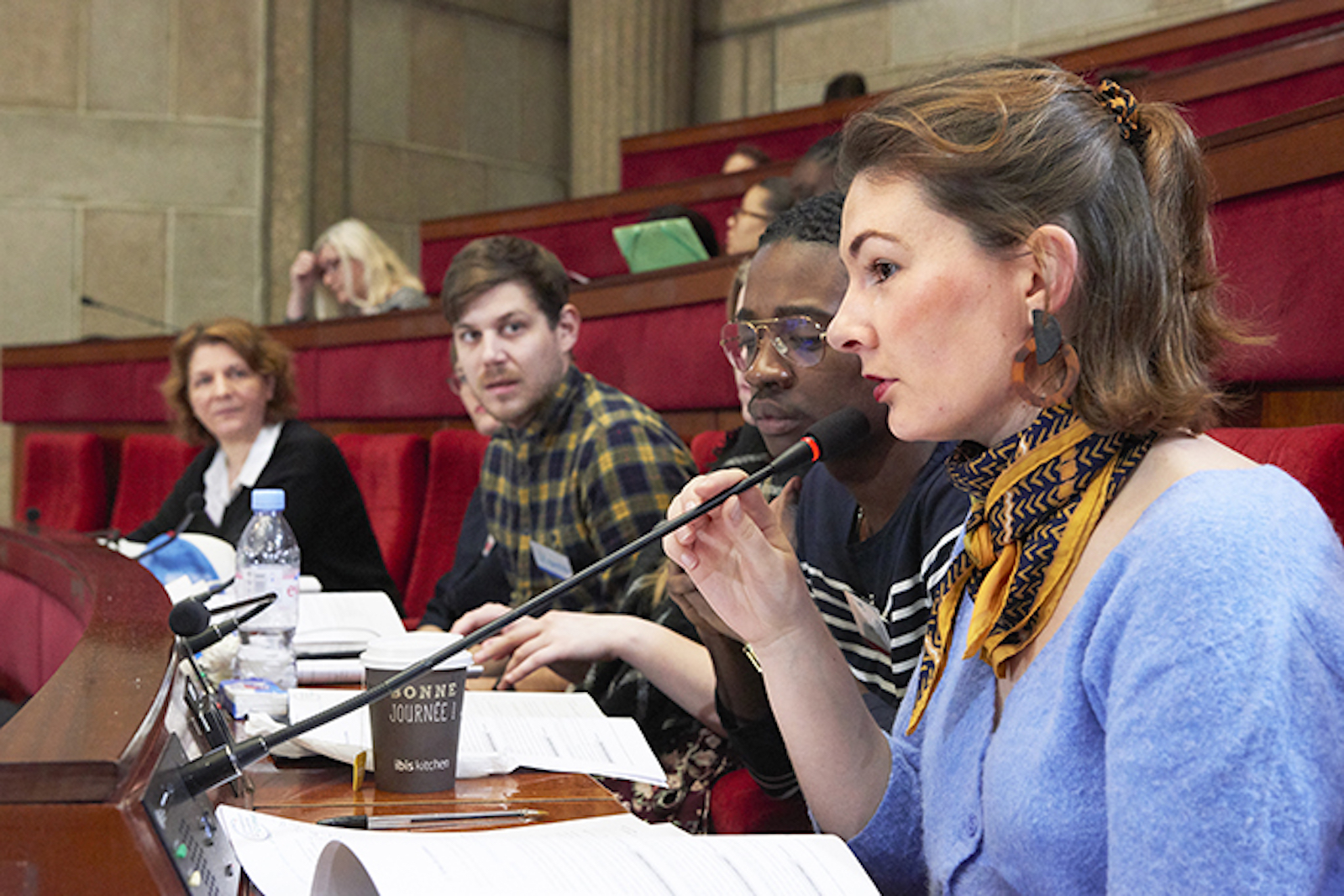 a woman in a blue sweater and yellow scarf holds a microphone as she speaks. She is seated next to several other people in red velvet chairs in a semicircular room