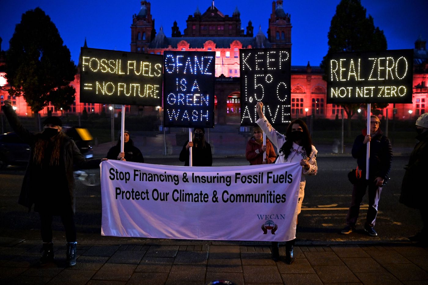 Protesters at night with signs that say "fossil fuels equal no futre" and "GFANZ is a green wash" and "keep 1.5 degrees alive" and "real zero not net zero"