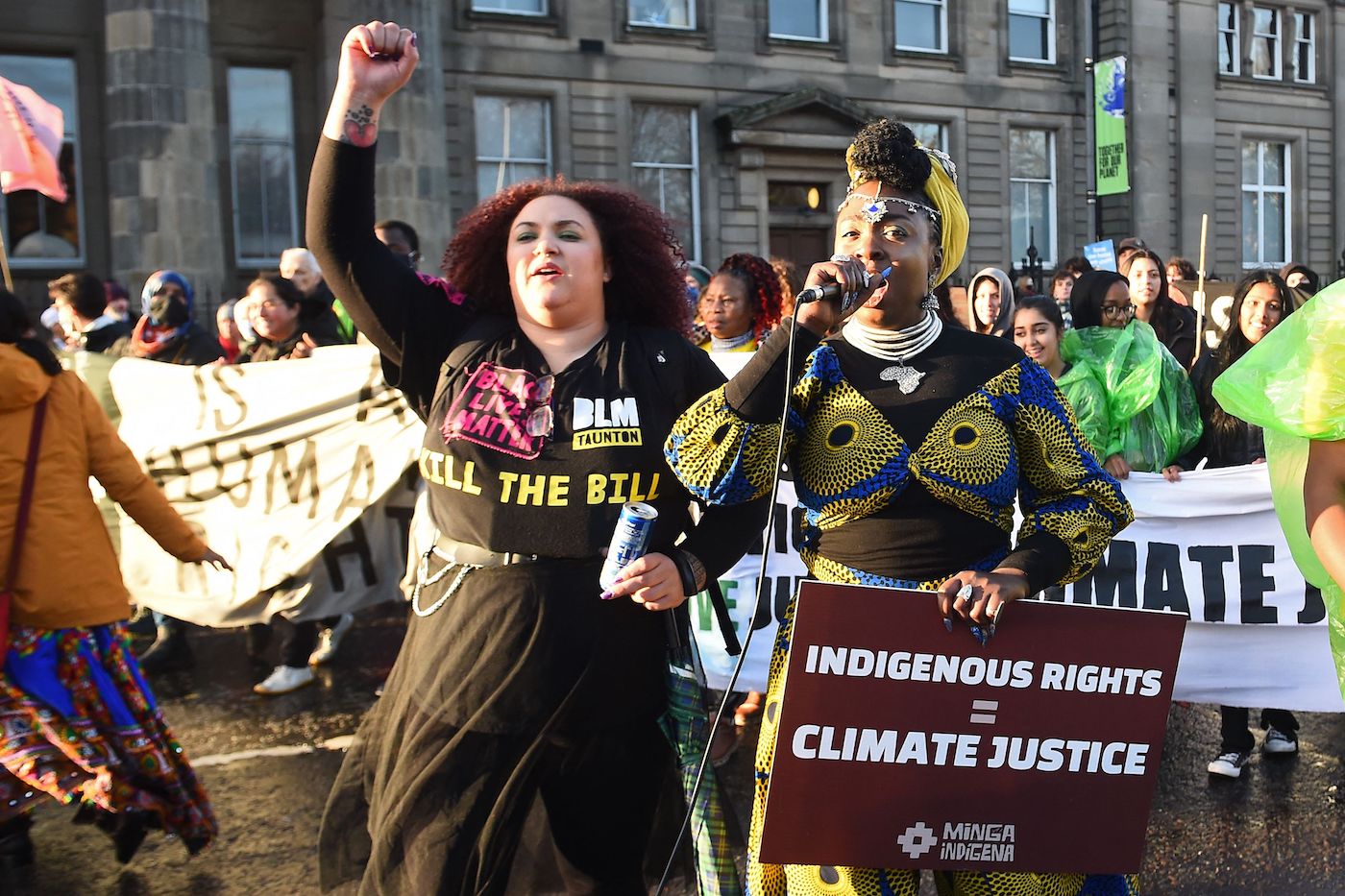 two women stand in protest. one woman has a black lives matter shirt on and the other is carrying a sign that says indigenous rights equal climate justice.