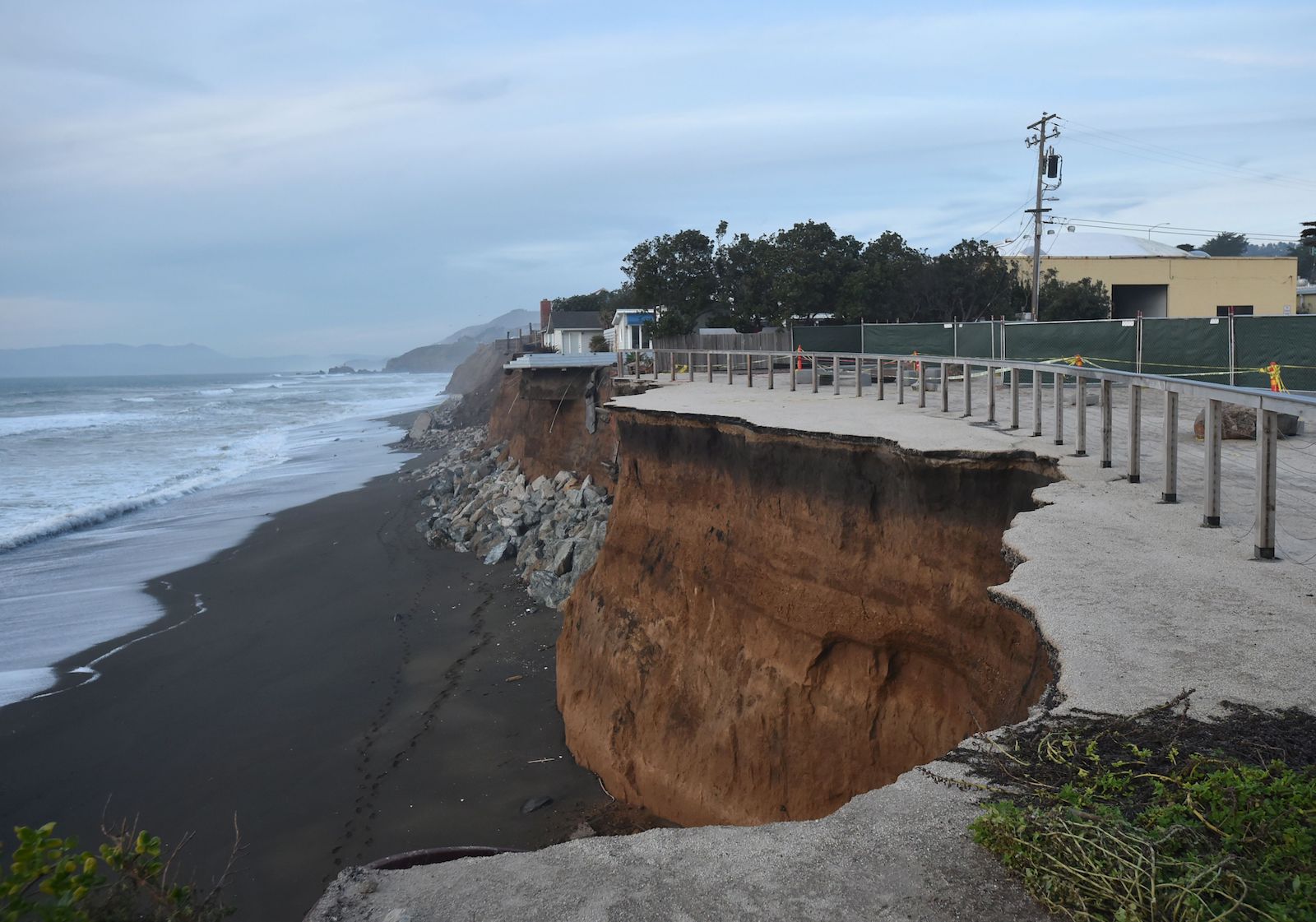 A high cliff-side road near the ocean shows signs of major breakage, with cracks coming right up the barrier of the road. Houses and powerlines are on the other side of the barrier on top of the cliff