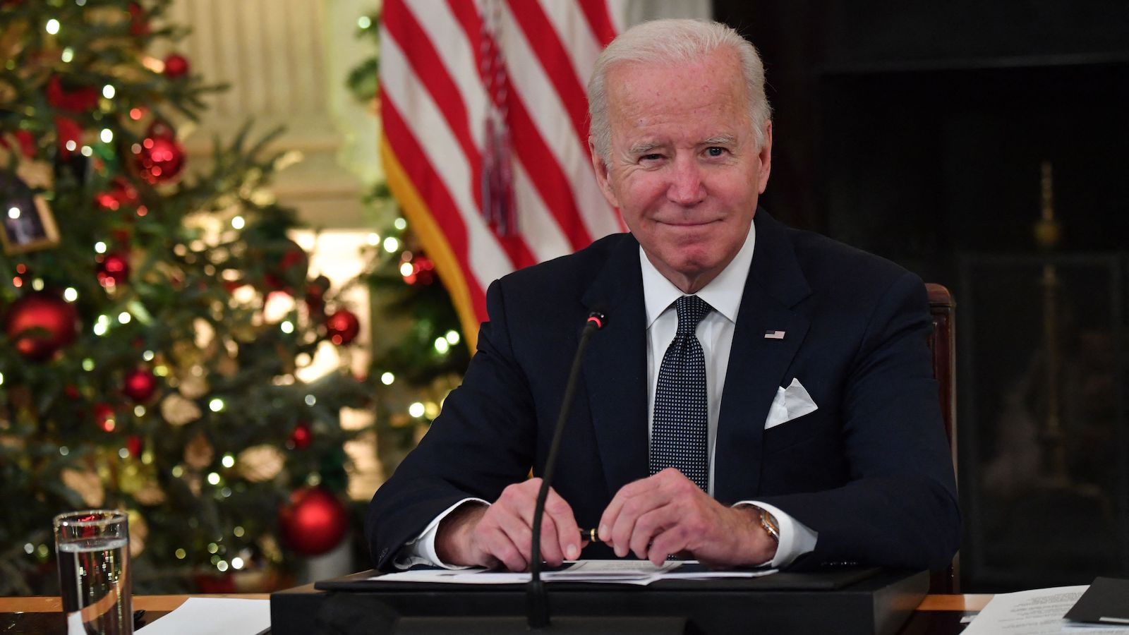 Joe Biden sitting at a desk with a christmas tree in the background with a wink on his face