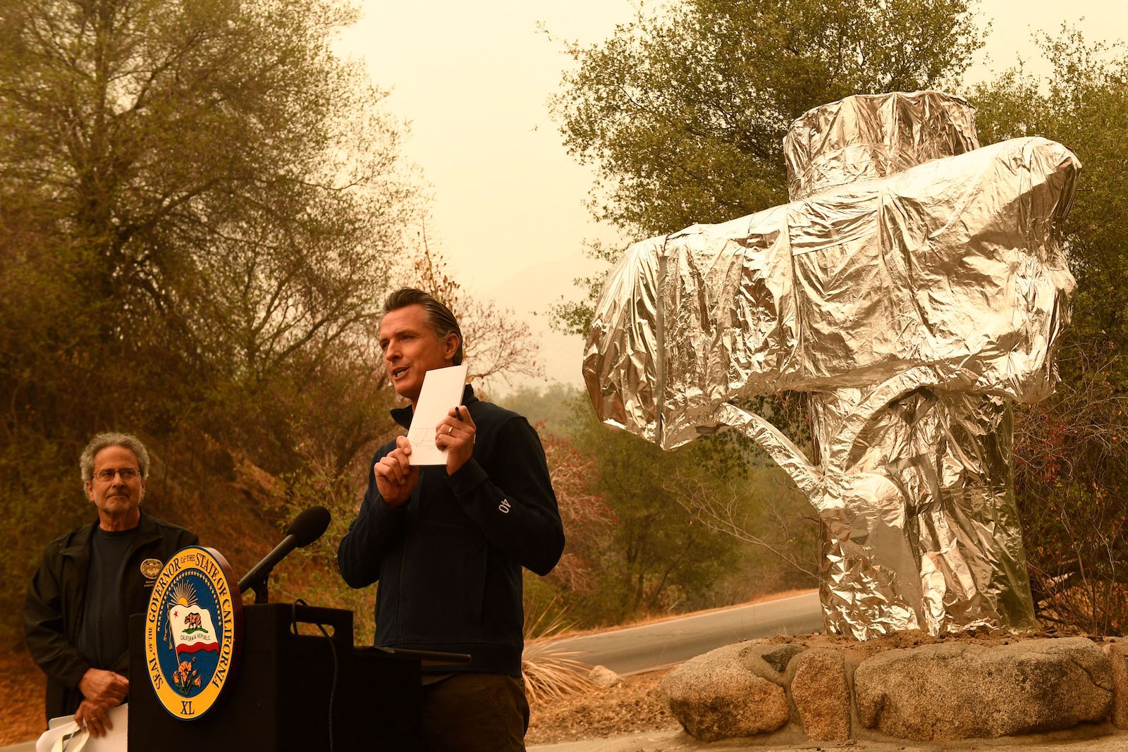 Gavin newsom holds white piece of paper while standing in front of podium. In the background, a foil-wrapped structure and trees and orange sky