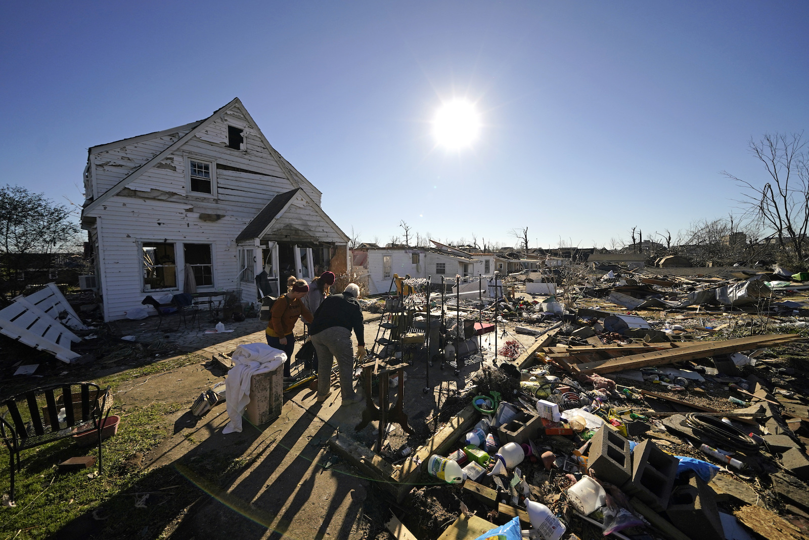 a white house with peeling paint and loose slats stands in front of a giant field of debris -- wood, bottles, and scrap metal -- through which people are walking. Overhead, a blazing sun looks down in a clear, blue sky.